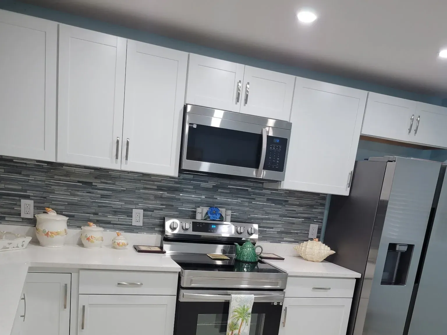 Finished kitchen with white shaker cabinets, blue-gray glass mosaic backsplash, white quartz counters and stainless appliances