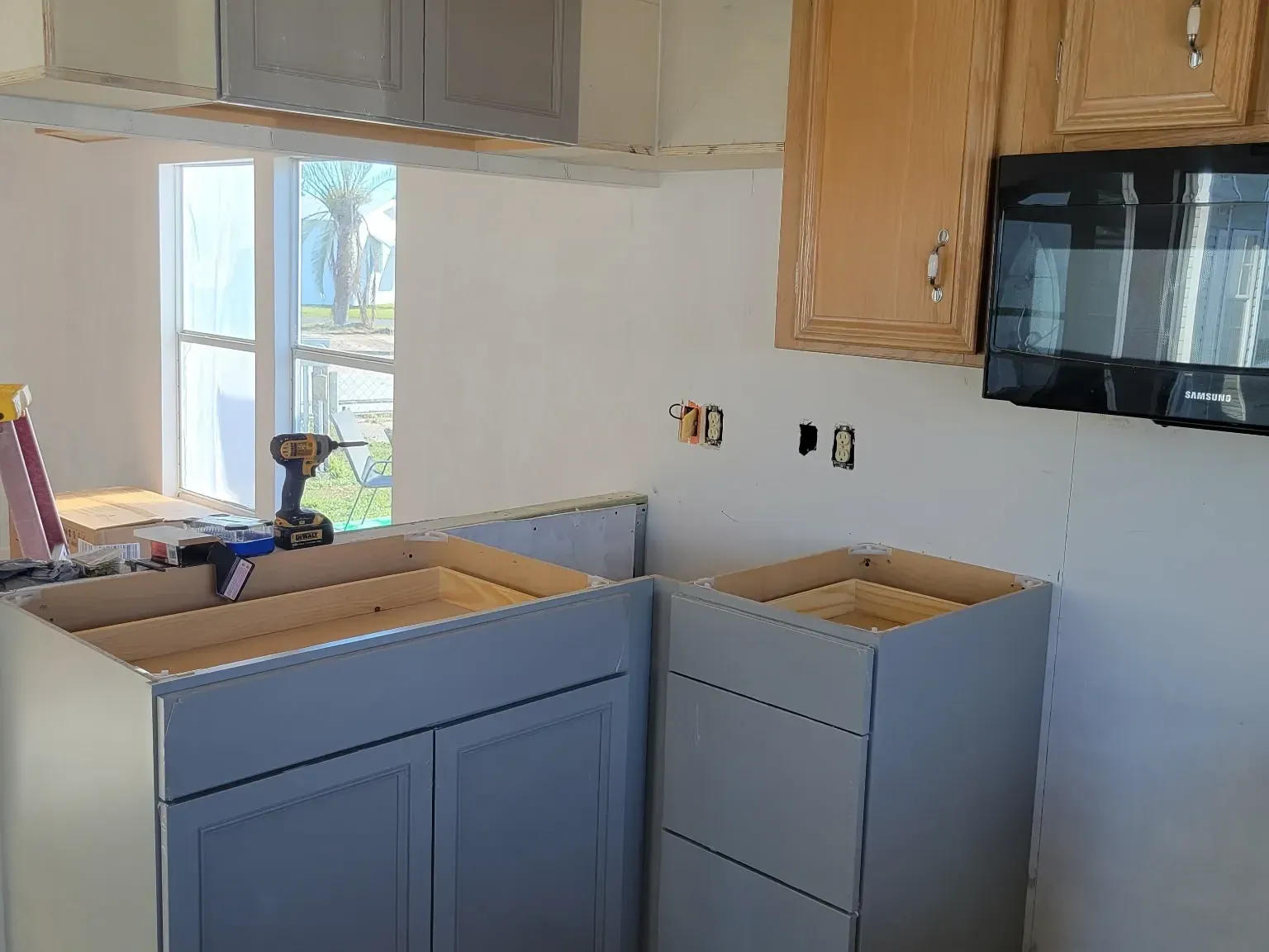 New gray base and wall cabinets set beside the original oak uppers, a cordless drill resting on an open cabinet box, outlet boxes exposed