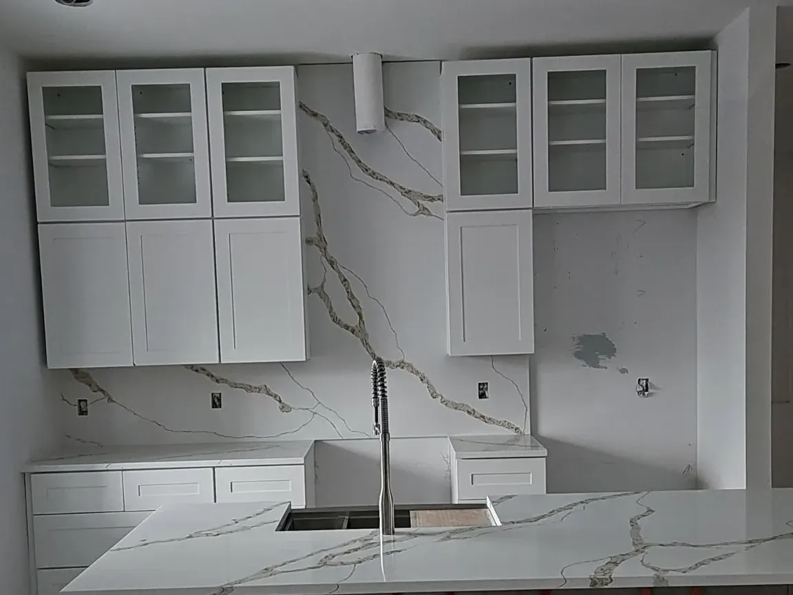 Kitchen mid-install with a Calacatta-look quartz slab backsplash, matching waterfall island with undermount sink and glass-front white uppers