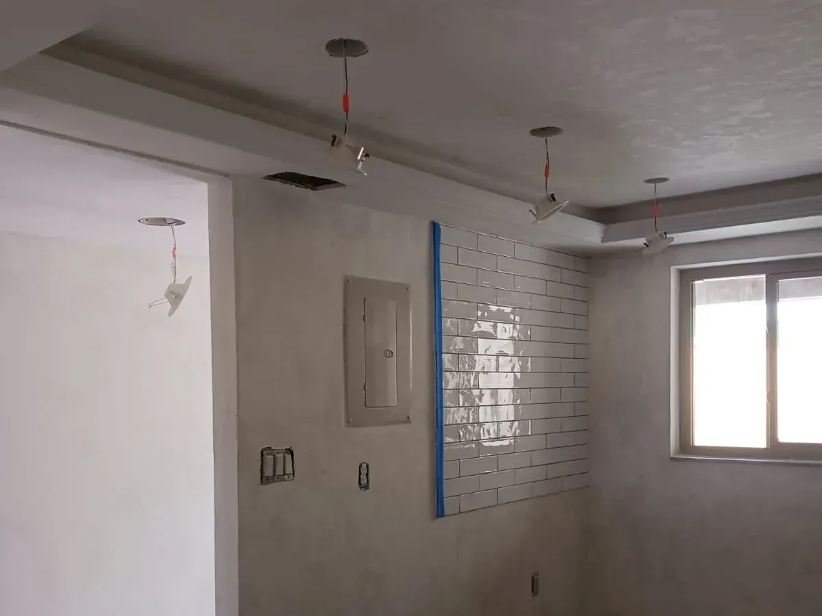 Recessed light housings hanging on their wiring above skim-coated walls, an open electrical panel and a taped-off section of new subway tile