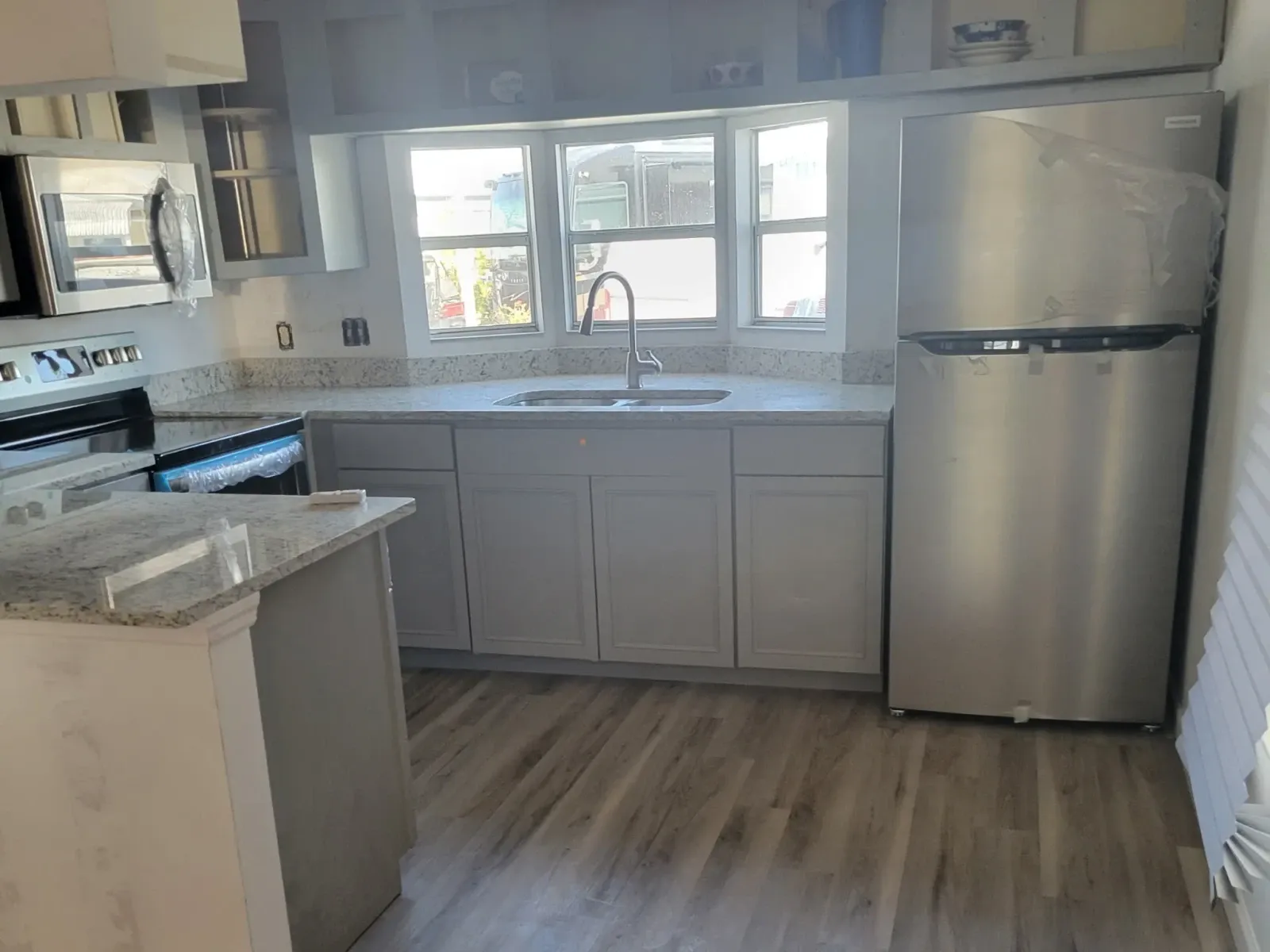 Completed gray shaker kitchen with speckled granite counters, black gooseneck faucet under a bay window, stainless appliances and gray plank flooring