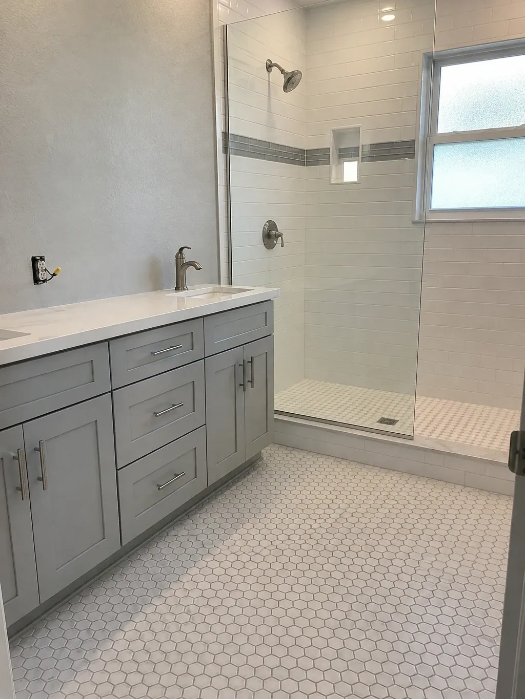 Gray shaker vanity with a quartz top, hexagon tile floor and a glass-panel shower with an accent band