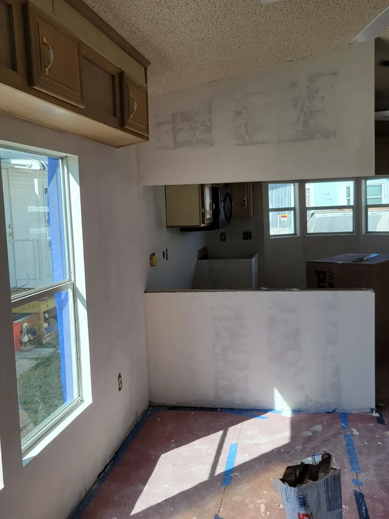 Open-plan kitchen and dining area mid-renovation with a newly drywalled half-wall, skim-coated walls and floor protection down