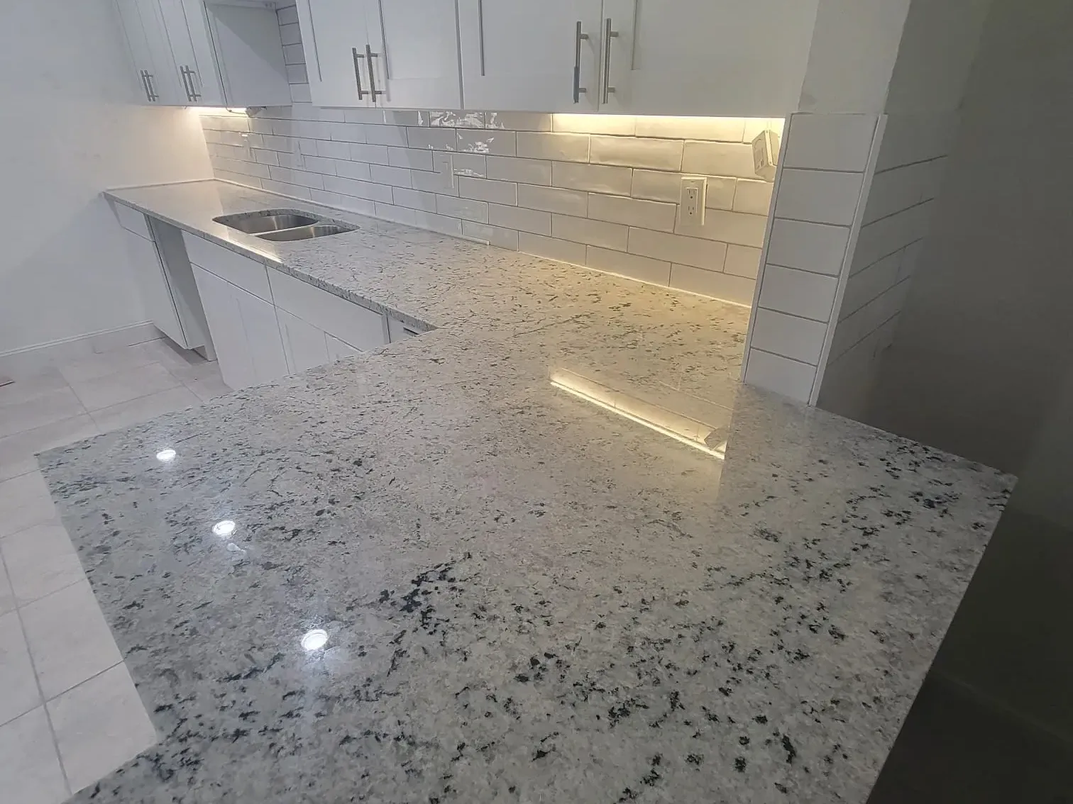 Galley kitchen with black-speckled white granite, stainless undermount sink and glossy subway tile lit by under-cabinet LED strips