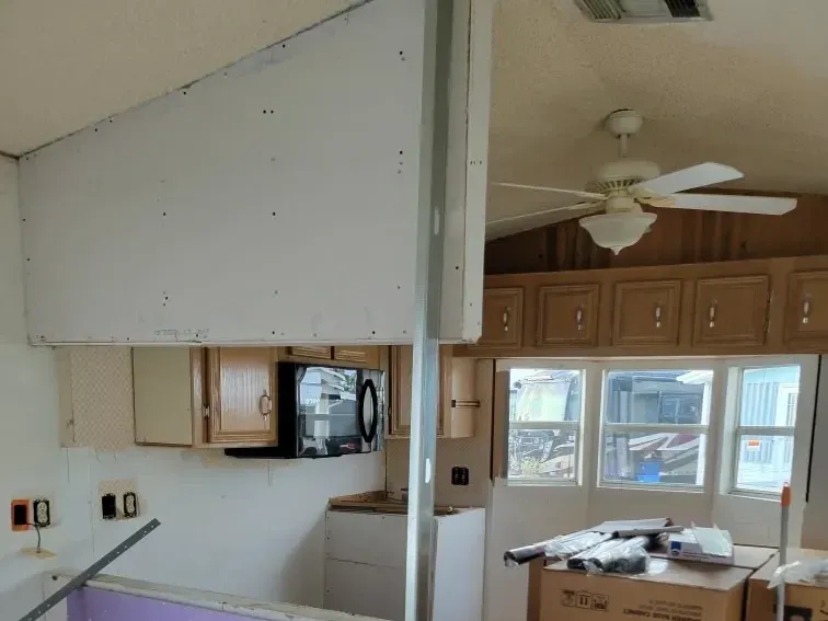 Kitchen soffit stripped to bare drywall with a temporary steel support post in place, original cabinets still hung and new cabinet cartons staged