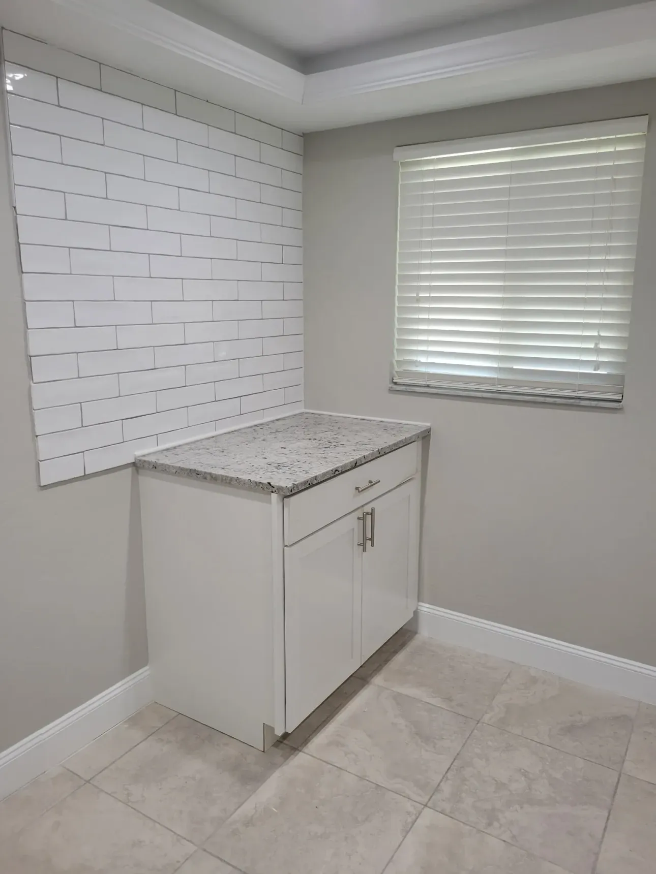Finished kitchen corner with white subway tile on two walls, a granite-topped white shaker cabinet and travertine-look floor tile