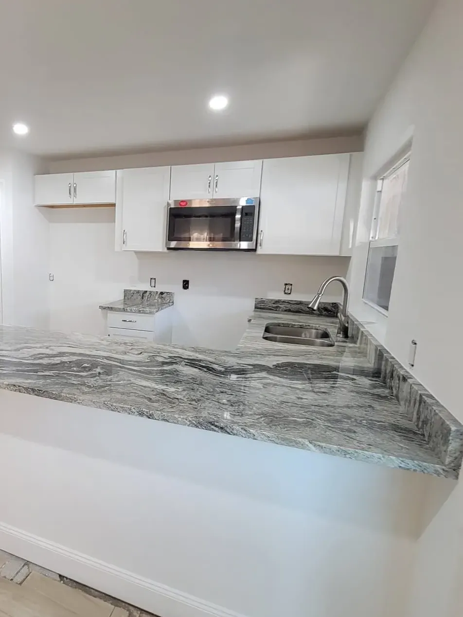Open kitchen with white shaker cabinets, a gray-veined granite peninsula, stainless microwave and wood-look plank tile floor