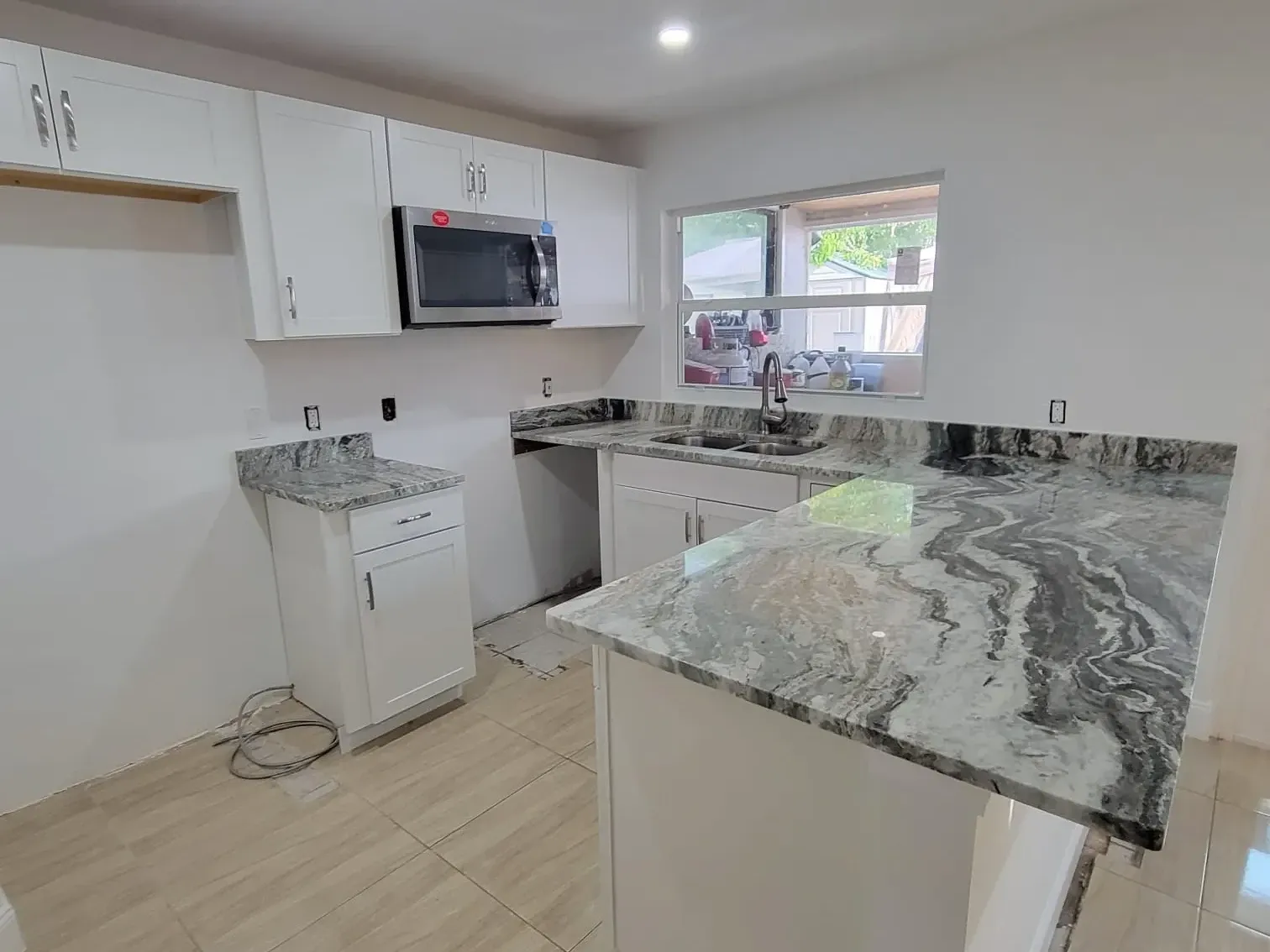 Heavily veined gray granite peninsula with white shaker cabinets, stainless microwave and a window over the double-bowl sink