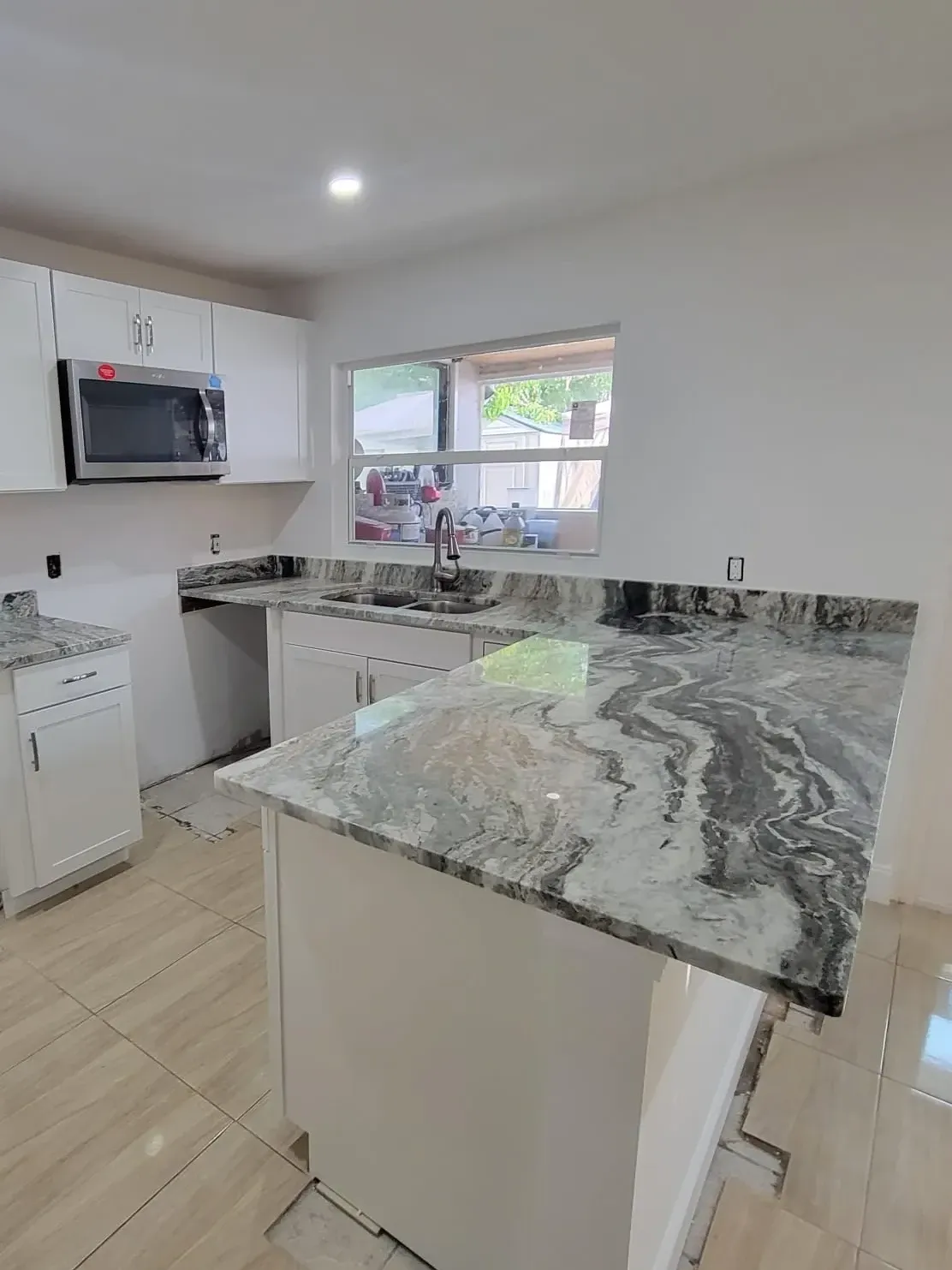 Heavily veined gray granite peninsula with white shaker cabinets, stainless microwave and a window over the double-bowl sink